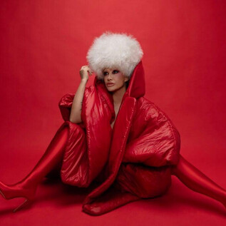 Žena sedí před červeným pozadím, oblečená v nápadném červeném plášti a vysokých červených botách. Nosí velkou bílou kožešinovou čepici a elegantně pózuje. | © Studio1538