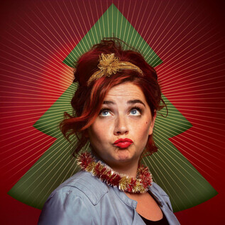 Eine Person posiert mit einem schrägen Gesichtsausdruck vor einem bunten Weihnachtsbaum-Hintergrund. Sie trägt eine festliche Halskette und eine Dekoration im Haar. | © Andrea Peller