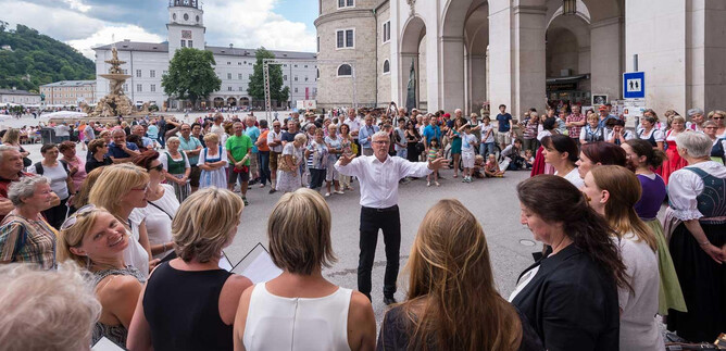 A large crowd gathers in a square to listen to a choir performance. In the foreground, a conductor leads the singers. | © Chorvernband Salzburg/Moser