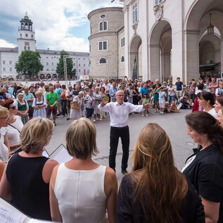 A large crowd gathers in a square to listen to a choir performance. In the foreground, a conductor leads the singers. | © Chorvernband Salzburg/Moser