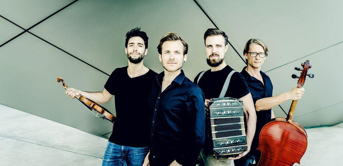 Un groupe de musique de quatre personnes se tient devant un mur moderne et géométrique. Chaque musicien tient un instrument, y compris un violon, un violoncelle et une autre machine musicale plus spécifique. | © Andrej Grilc