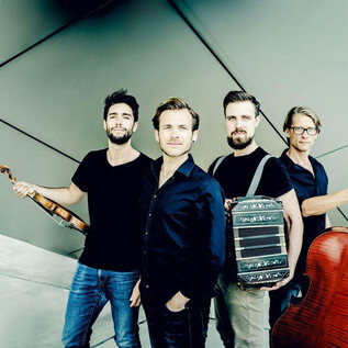 Un groupe de musique de quatre personnes se tient devant un mur moderne et géométrique. Chaque musicien tient un instrument, y compris un violon, un violoncelle et une autre machine musicale plus spécifique. | © Andrej Grilc