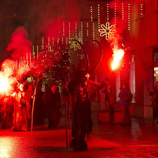 Eine Menschenmenge mit bengalischen Fackeln feiert in einer festlich beleuchteten Straße. Rote Lichter schaffen eine lebhafte und festliche Atmosphäre. | © Sternadvent