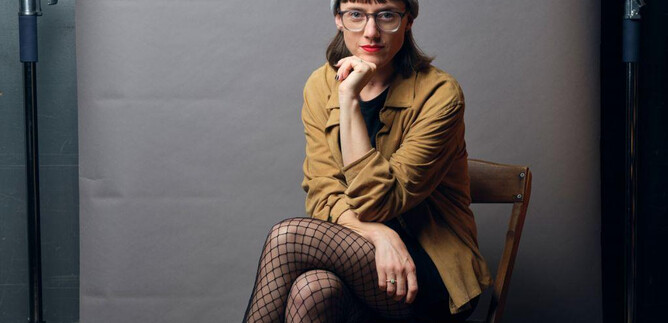 A person is sitting on a chair in front of a gray background. They are wearing a gray beanie, a brown jacket, and fishnet tights. | © Remo Büss