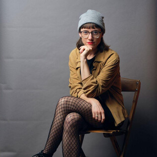 A person is sitting on a chair in front of a gray background. They are wearing a gray beanie, a brown jacket, and fishnet tights. | © Remo Büss