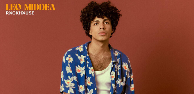 Ein junger Mann mit lockigem Haar trägt ein buntes, blumiges Hemd. Der Hintergrund ist in einem warmen Farbton gehalten. | © Elisa Maciel