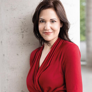 Žena v černém obleku a s kudrnatými vlasy stojí s úsměvem před oranžovou zdí. Má zkřížené ruce a působí přátelsky. | © Alexandra Grill