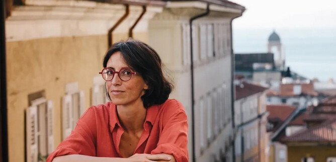 Eine Frau mit schwarzem Anzug und lockigem Haar steht lächelnd vor einer orangefarbenen Wand. Sie hat die Arme verschränkt und wirkt freundlich. | © Adolfo Frediani