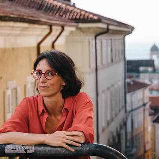 Une femme en costume noir et avec des cheveux bouclés se tient souriante devant un mur orange. Elle a les bras croisés et a l'air amical. | © Adolfo Frediani