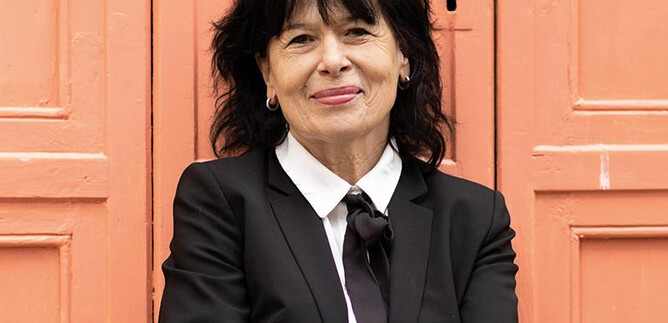 Eine Frau mit schwarzem Anzug und lockigem Haar steht lächelnd vor einer orangefarbenen Wand. Sie hat die Arme verschränkt und wirkt freundlich. | © Minitta Kandlbauer