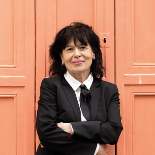 Une femme en costume noir et avec des cheveux bouclés se tient souriante devant un mur orange. Elle a les bras croisés et a l'air amical. | © Minitta Kandlbauer