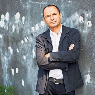 Žena v černém obleku a s kudrnatými vlasy stojí s úsměvem před oranžovou zdí. Má zkřížené ruce a působí přátelsky. | © Gunter Gluecklich