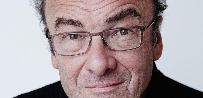 Eine Frau mit schwarzem Anzug und lockigem Haar steht lächelnd vor einer orangefarbenen Wand. Sie hat die Arme verschränkt und wirkt freundlich. | © Rafaela Proell