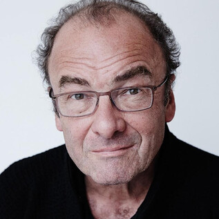 Une femme en costume noir et avec des cheveux bouclés se tient souriante devant un mur orange. Elle a les bras croisés et a l'air amical. | © Rafaela Proell