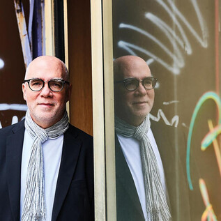 Žena v černém obleku a s kudrnatými vlasy stojí s úsměvem před oranžovou zdí. Má zkřížené ruce a působí přátelsky. | © Aleksandra Pawloff