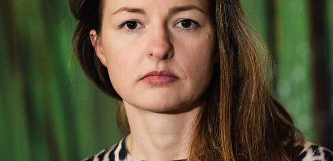 Žena v černém obleku a s kudrnatými vlasy stojí s úsměvem před oranžovou zdí. Má zkřížené ruce a působí přátelsky. | © Clemens Schmiedbauer