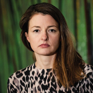 A woman in a black suit with curly hair stands smiling in front of an orange wall. She has her arms crossed and looks friendly. | © Clemens Schmiedbauer