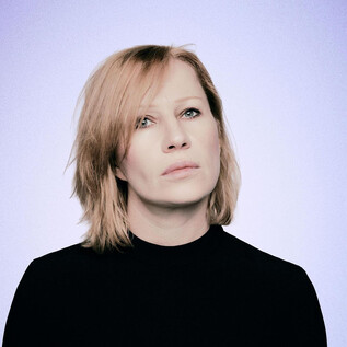 Une personne aux cheveux blonds légèrement en désordre regarde pensivement au loin. Elle porte un haut noir devant un arrière-plan doux et clair. | © Susanne Hassler-Smith