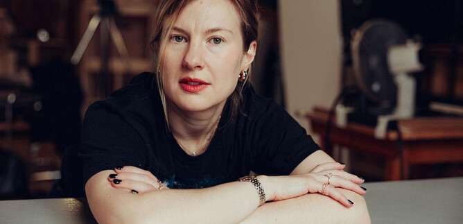 A woman is sitting at a table and looking directly at the camera. She is wearing a black T-shirt and has a thoughtful expression. | © Joel Heyd