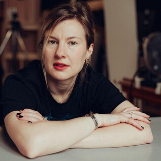 Une femme est assise à une table et regarde directement la caméra. Elle porte un T-shirt noir et a une expression pensive. | © Joel Heyd