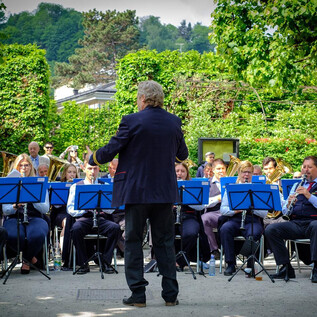 Un ensemble de cuivres joue en plein air. Le chef d'orchestre se tient devant le groupe, entouré d'arbres verts. | © knaro.at