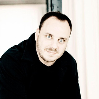 Un homme aux cheveux courts se tient dans une pièce claire. Il a une expression amicale et porte un haut noir. | © Marco Borggreve