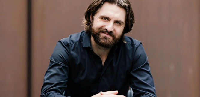 A man with a beard is sitting at a table and looking kindly at the camera. He is wearing a black shirt and has long, wavy hair. | © Geoffroy_Schied
