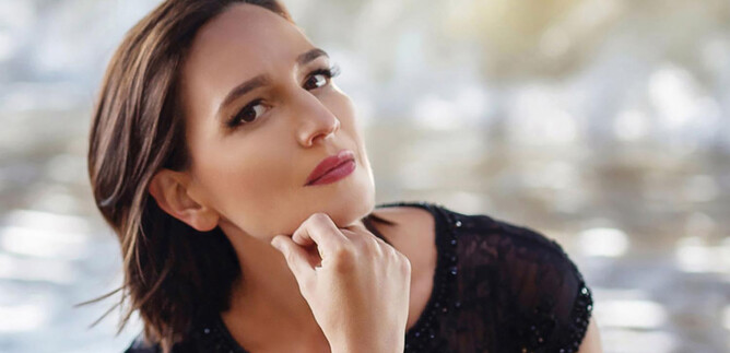 Žena s krátkými hnědými vlasy sedí zamyšleně u vody. Na sobě má černý top a dívá se přímo do kamery. | © Yulia Oliver-Taylor