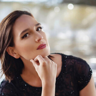 A woman with short, brown hair sits thoughtfully by the water. She is wearing a black top and looks directly into the camera. | © Yulia Oliver-Taylor