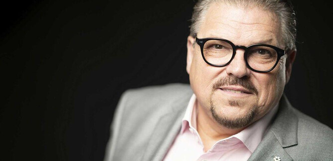 Un homme avec des lunettes et une apparence soignée sourit légèrement. Il porte un costume gris et une chemise claire. | © Lukas Beck