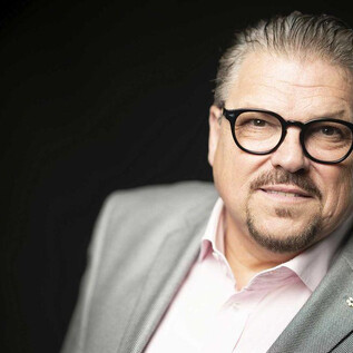 A man with glasses and a well-groomed appearance smiles slightly. He is wearing a gray suit and a light-colored shirt. | © Lukas Beck