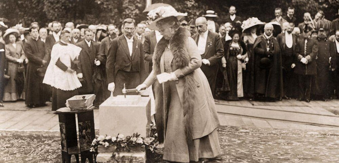 Une femme en vêtements élégants pose une pierre sur une table, tandis qu'une grande foule regarde en arrière-plan. La scène se déroule manifestement lors d'un événement ou d'une cérémonie solennelle. | © ISM
