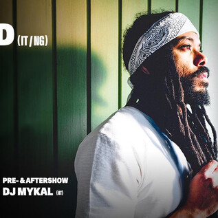 Ein Mann mit langen, lockigen Haaren steht in einem leicht schattigen Bereich. Er trägt ein weißes Oberteil und schaut nachdenklich zur Seite. | © Rockhouse Salzburg