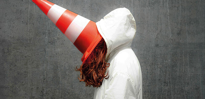 Une personne porte une veste blanche et a un cône de signalisation sur la tête. L'arrière-plan est gris et simple. | © Valentina Schuster