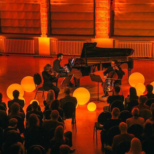 A live music performance featuring three musicians on stage. The space is surrounded by warm light and decorative balls, while the audience follows the performance. | © Freie Kunst