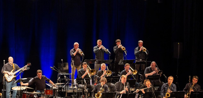 Eine große Band spielt auf der Bühne. Im Hintergrund sind Musiker mit Instrumenten und eine blaue Bühnenbeleuchtung zu sehen. | © Albert Moser