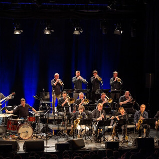 A large band is playing on stage. In the background, musicians with instruments and blue stage lighting can be seen. | © Albert Moser