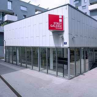 A modern building with large window areas and a red sign. It is the city gallery, located in an urban environment. | © Hermann Seidl