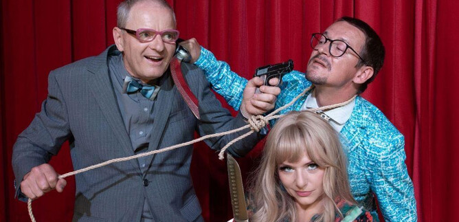 Drei Personen in einem komischen Szenario, wobei einer eine Waffe hält und die andere mit einem Seil gebunden ist. Im Hintergrund ist ein roter Vorhang zu sehen. | © Sigrid Riepl