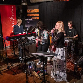 Una banda toca en vivo en el escenario de un club de música. Los miembros están compuestos por diferentes instrumentistas y una cantante. | © Musikum