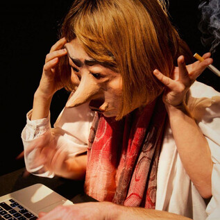 Eine Person mit einer großen Maske sitzt vor einem Laptop. Sie sieht gestresst aus und hält eine Zigarette in der Hand. | © Debra Tchotchov