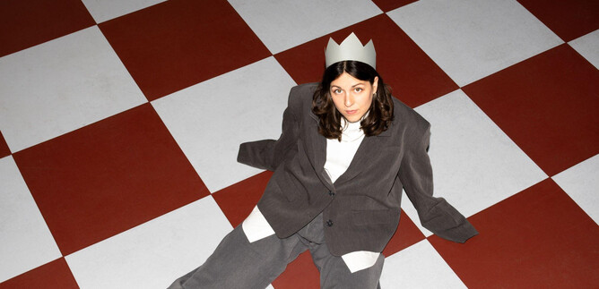 A person is sitting on a chessboard floor wearing a crown hat. They are dressed in a gray suit and looking directly at the camera. | © Sima Prodinger