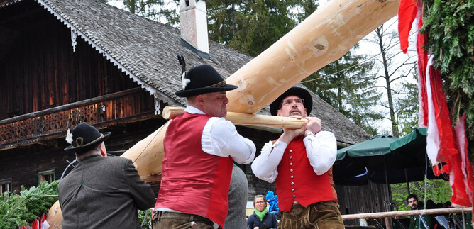 Men in traditional costumes are lifting a heavy wooden beam. In the background, a rustic house and nature can be seen. | © Salzburger Freilichtmuseum
