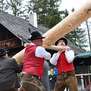 Männer in traditionellen Trachten heben einen schweren Holzträger. Im Hintergrund ist ein rustikales Haus und die Natur zu sehen. | © Salzburger Freilichtmuseum