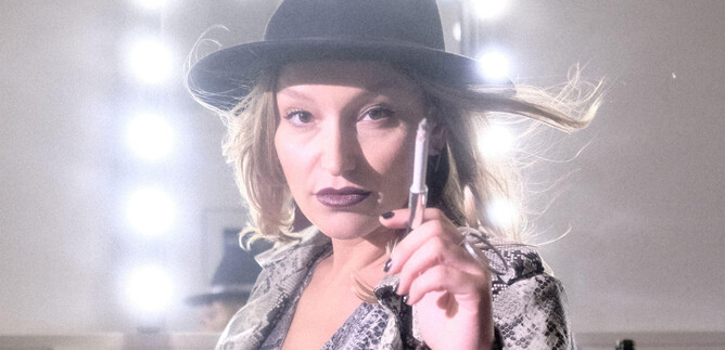 Una donna con un cappello e un'espressione sicura di sé sta davanti a uno specchio illuminato. Tiene una penna in mano e ha un look alla moda. | © Christopher Glanzl