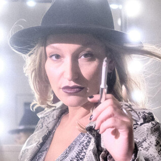 A woman with a hat and a confident expression stands in front of a lit mirror. She holds a pen in her hand and has a fashionable look. | © Christopher Glanzl