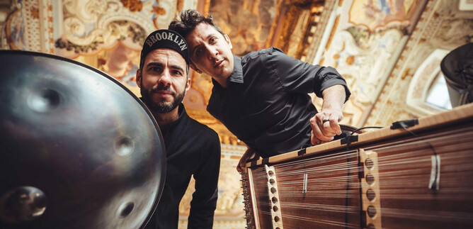 Two musicians pose with their instruments in an intricately decorated room. The background features magnificent walls and ceilings. | © Simon Reithofer