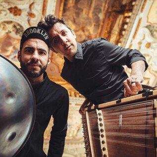 Two musicians pose with their instruments in an intricately decorated room. The background features magnificent walls and ceilings. | © Simon Reithofer