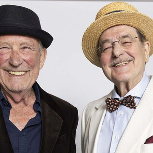 Dva starší muži se šťastně usmívají do kamery. První má na sobě černý klobouk, druhý má na sobě světlý slaměný klobouk a elegantní sako. | © Xenia Trampusch