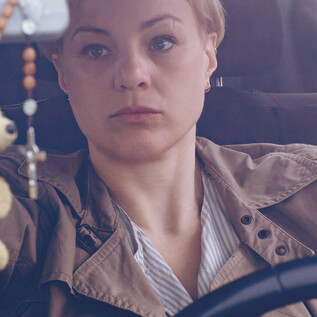 Egy nő ül az autó volánjánál és gondolkodva néz. A háttérben egy autós ülés és egy akasztott kulcstartó látható. | © Apollonia Theresa Bitzan